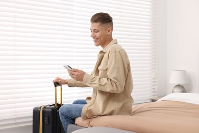 Photo of Smiling guest with suitcase and smartphone on bed in stylish hotel room