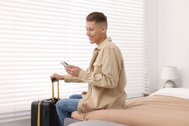 Smiling guest with suitcase and smartphone on bed in stylish hotel room