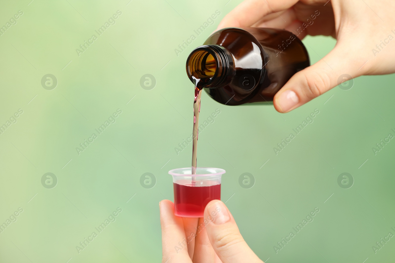 Photo of Woman pouring cough syrup into measuring cup on light green background, closeup