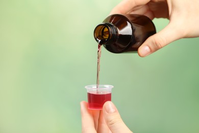 Photo of Woman pouring cough syrup into measuring cup on light green background, closeup