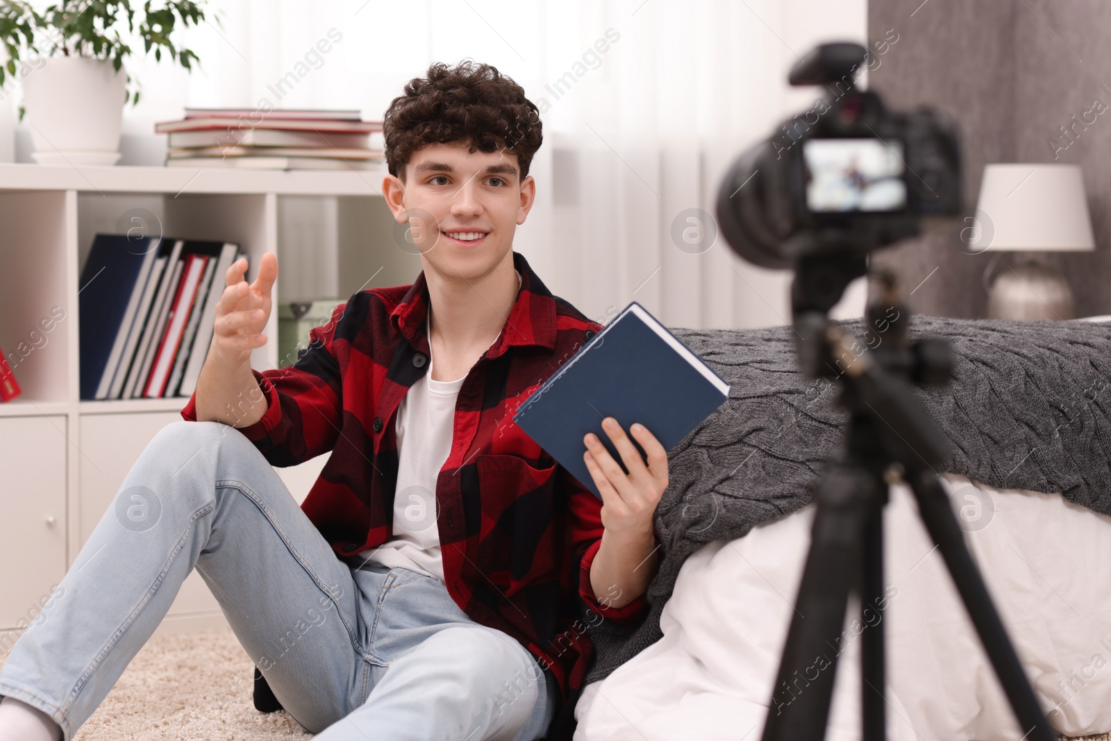 Photo of Smiling teenage blogger with book explaining something while streaming at home