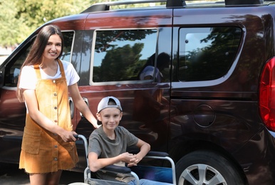 Photo of Young woman with boy in wheelchair near van outdoors
