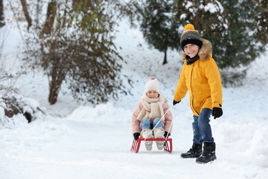 Little boy pulling sledge with his sister through snow in winter park, space for text