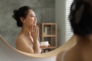 Happy woman applying face cream near mirror at home