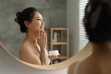 Photo of Happy woman applying face cream near mirror at home