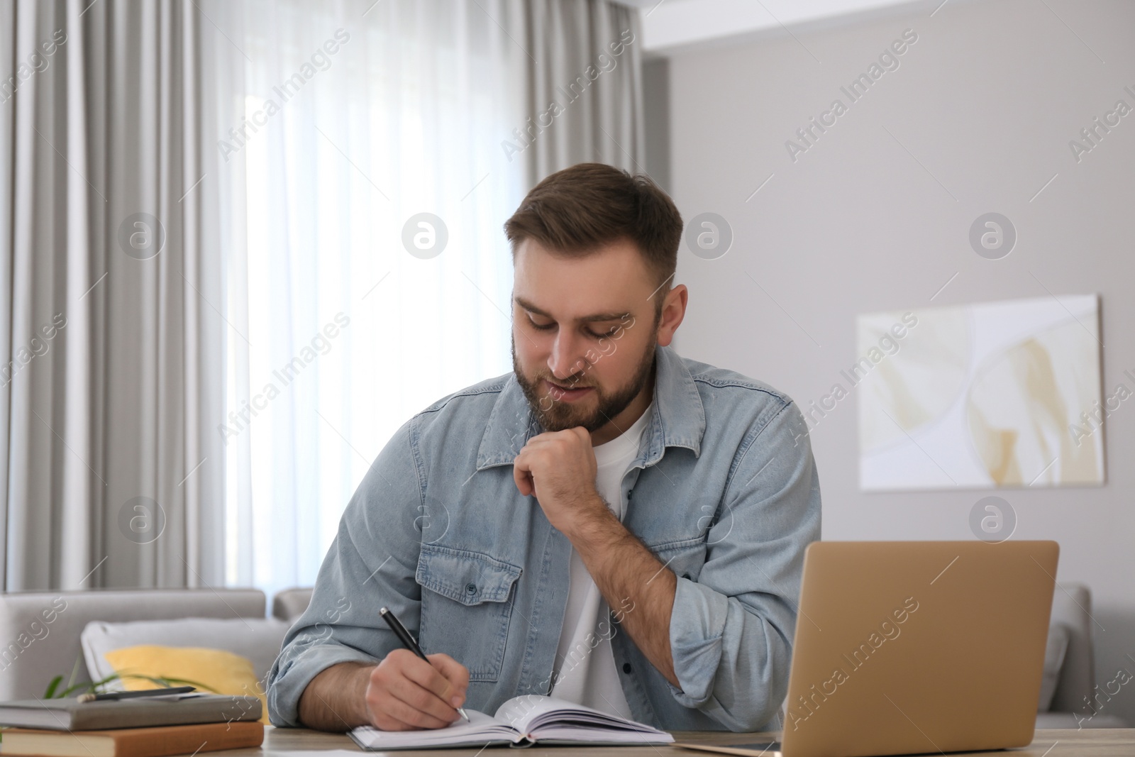 Photo of Young man taking notes during online webinar at table indoors