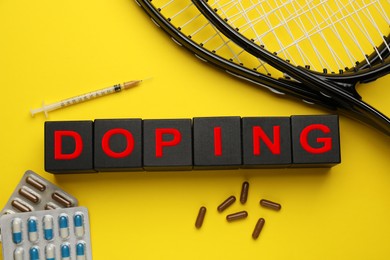 Photo of Black wooden cubes with word Doping, tennis rackets and drugs on yellow background, flat lay
