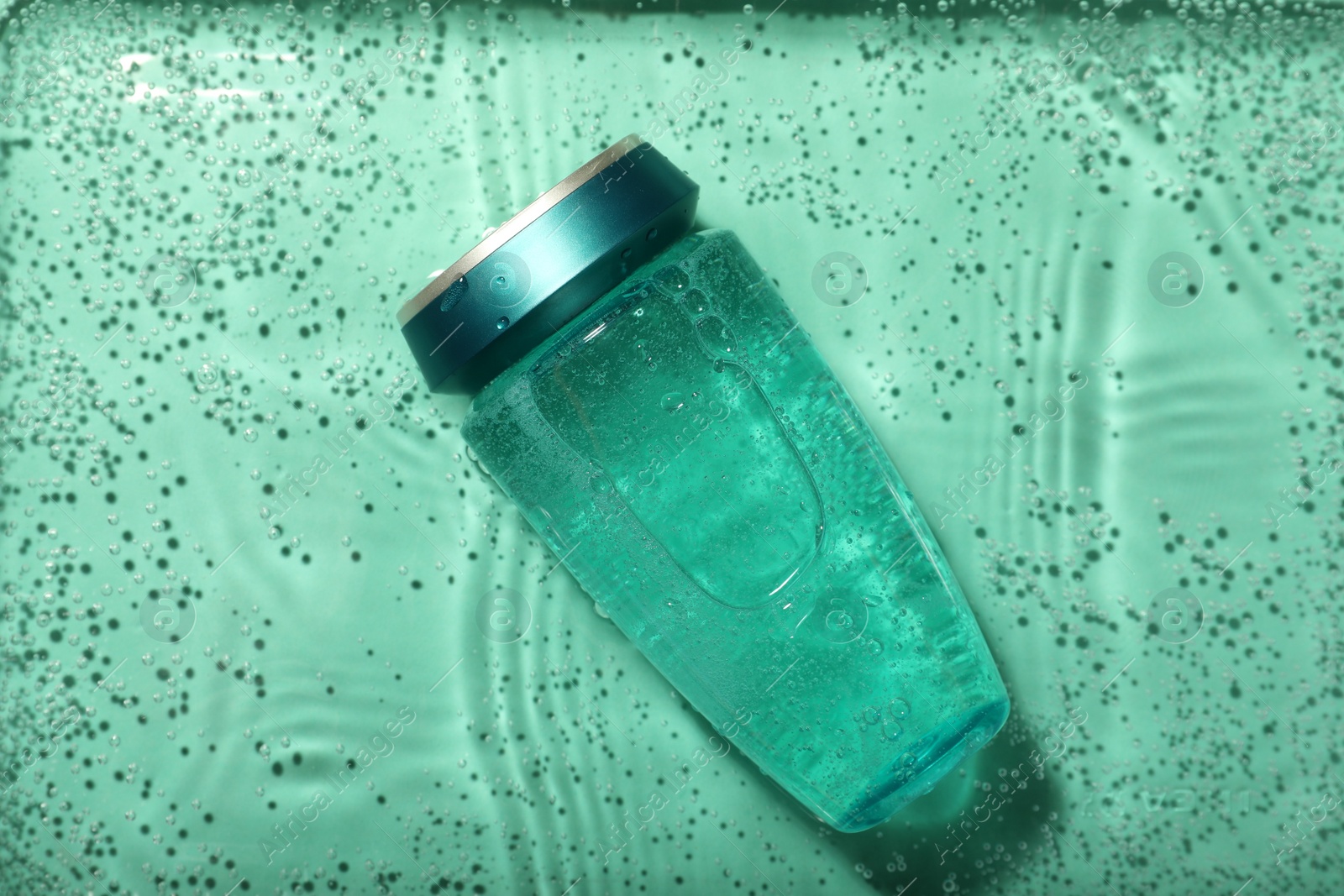 Photo of Bottle of hair care cosmetic product in water on turquoise background, top view