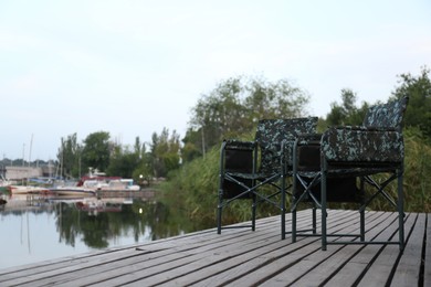 Photo of Camouflage fishing chairs on wooden pier near river, space for text