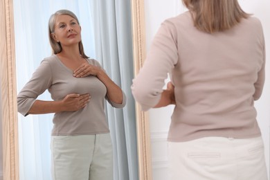 Photo of Beautiful senior woman doing breast self-examination near mirror indoors