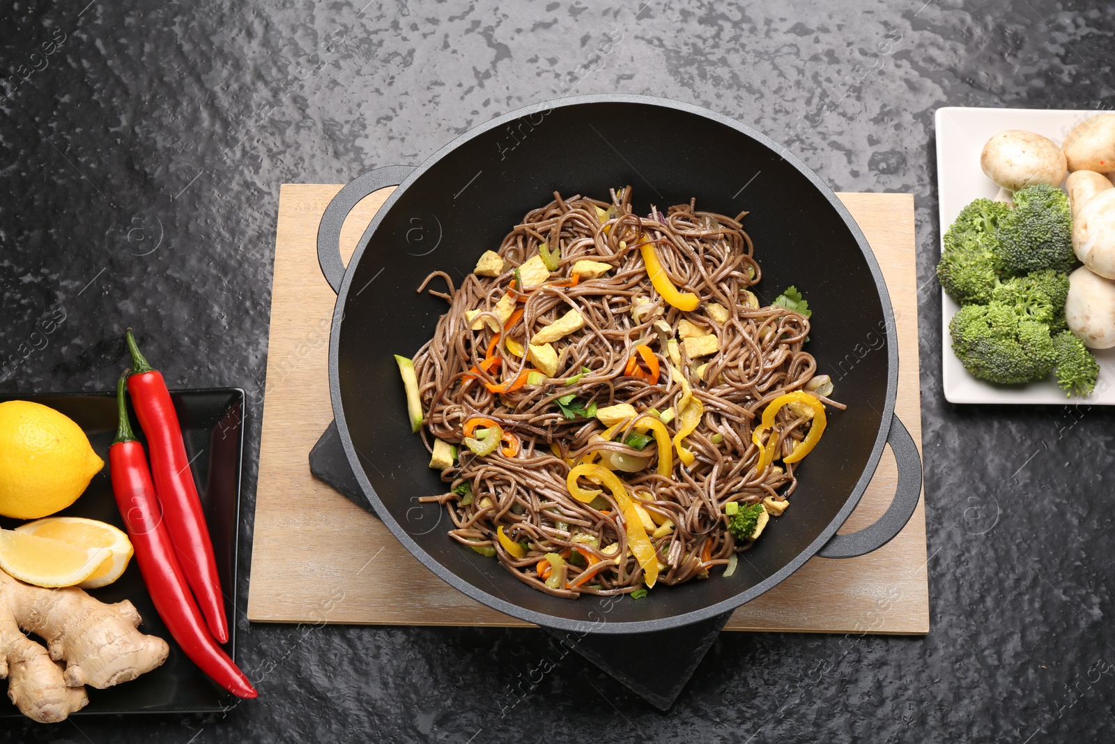 Photo of Stir-fry. Tasty noodles with vegetables, meat in wok and ingredients on dark textured table, flat lay