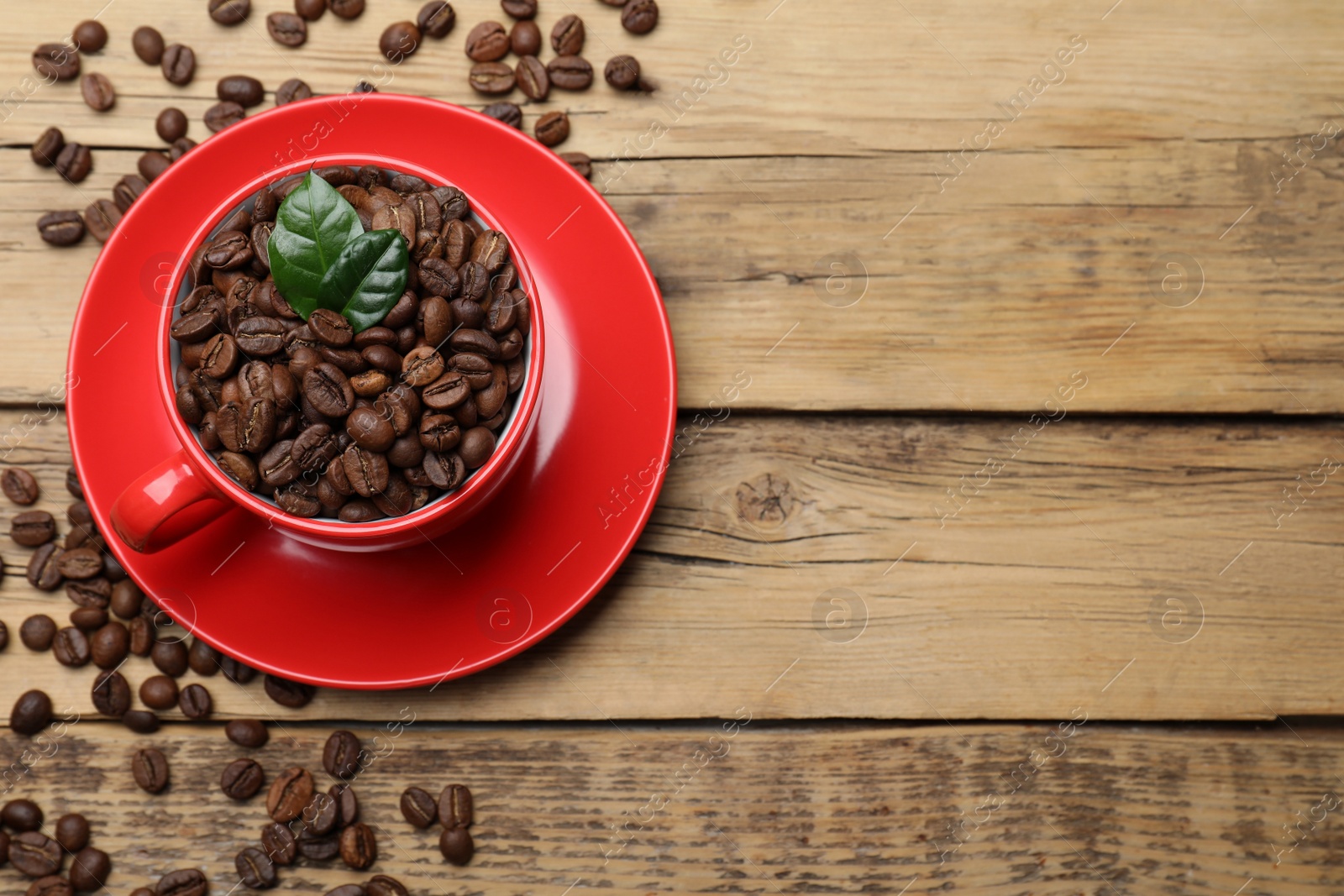 Photo of Red cup with roasted coffee beans and leaves on wooden table, flat lay. Space for text