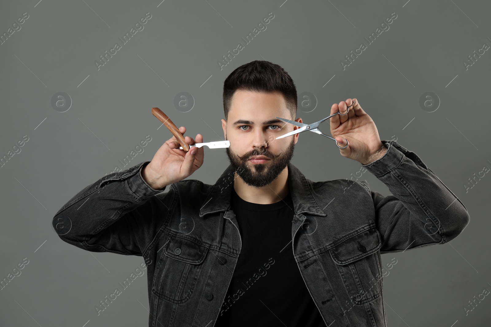 Photo of Handsome young man with mustache holding blade and scissors on grey background