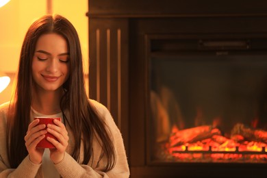 Photo of Young woman with cup of hot drink near fireplace indoors. Cozy atmosphere