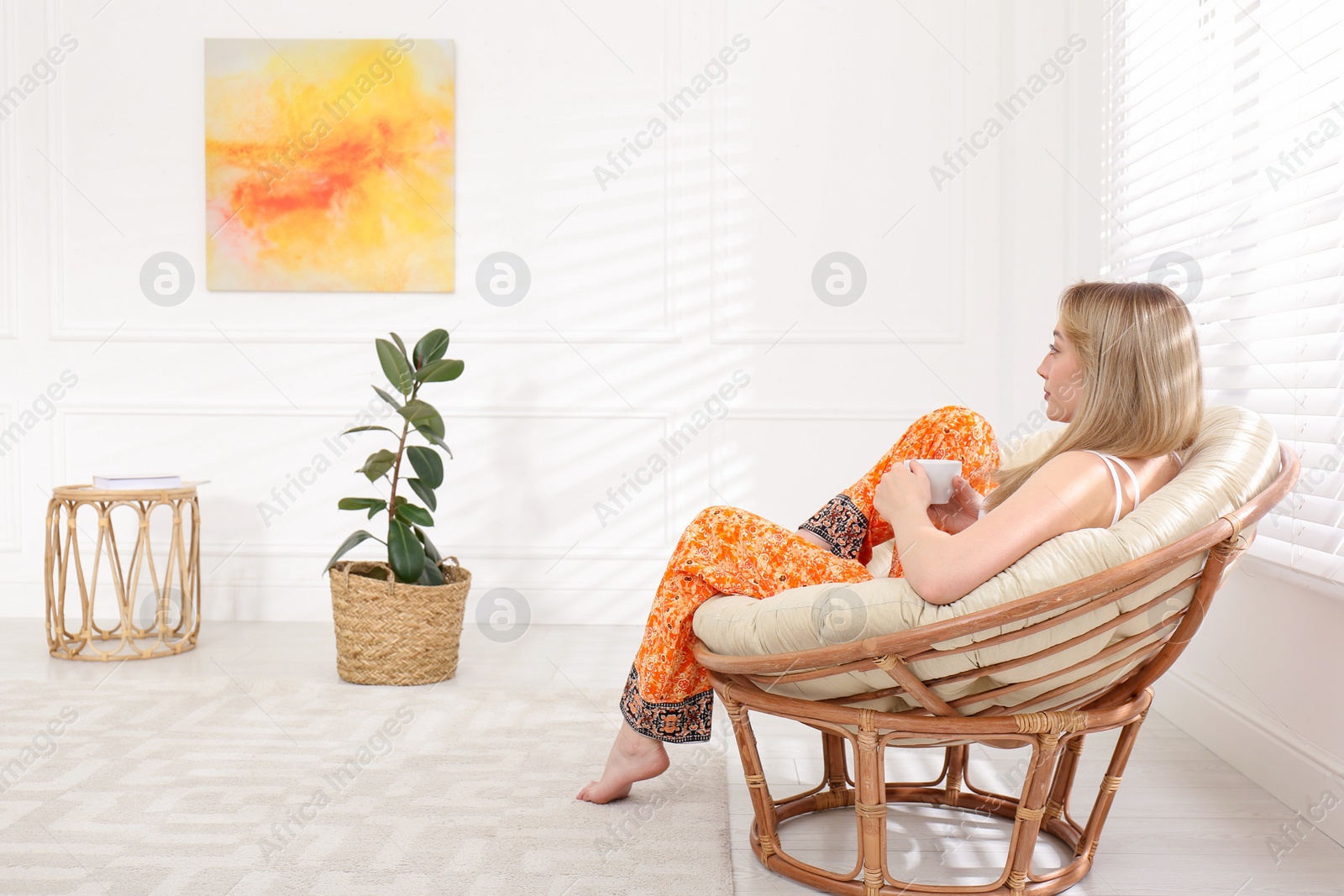 Photo of Beautiful young woman with cup of hot drink relaxing in papasan chair at home, space for text