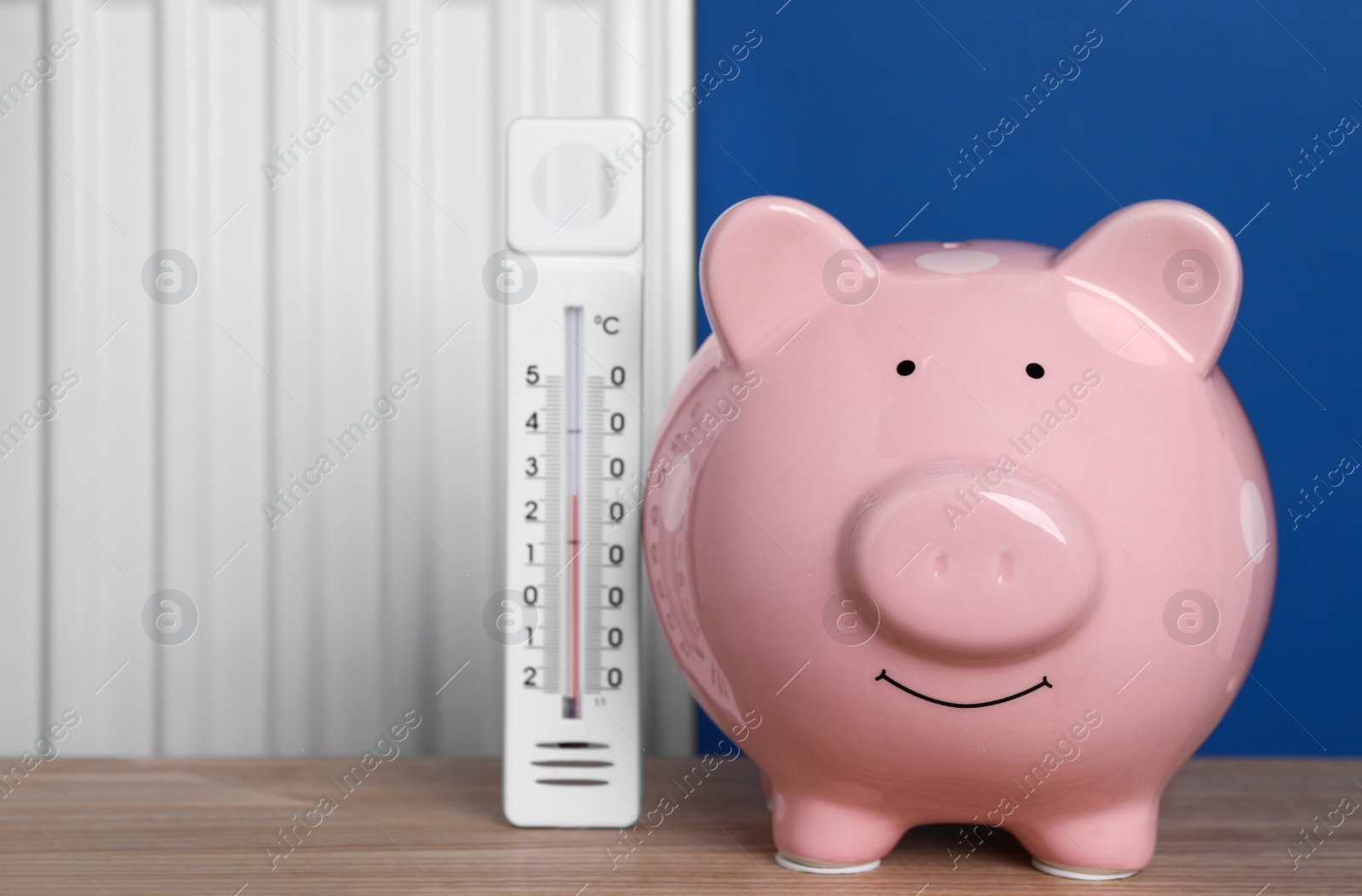 Photo of Piggy bank with thermometer on wooden table near heating radiator. Space for text