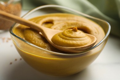 Photo of Spoon and glass bowl with tasty mustard sauce on white table, closeup