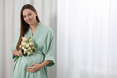 Photo of Beautiful pregnant woman with bouquet of roses near window indoors, space for text