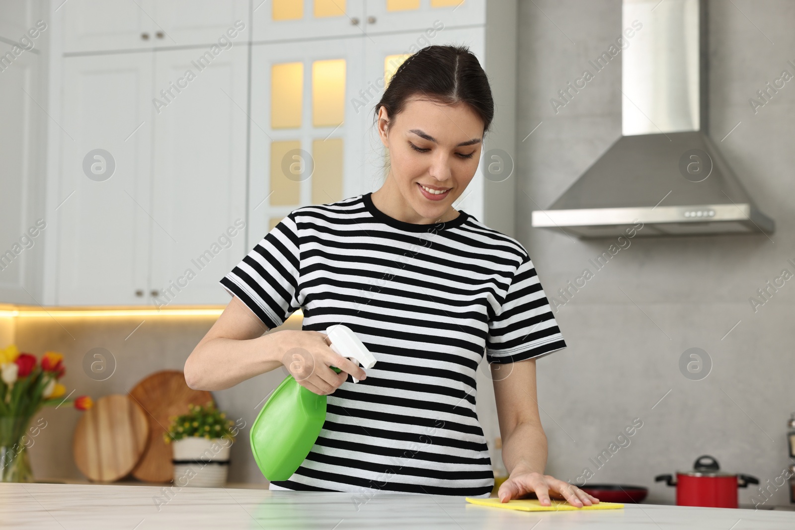 Photo of Woman cleaning countertop with sponge wipe and spray bottle in kitchen