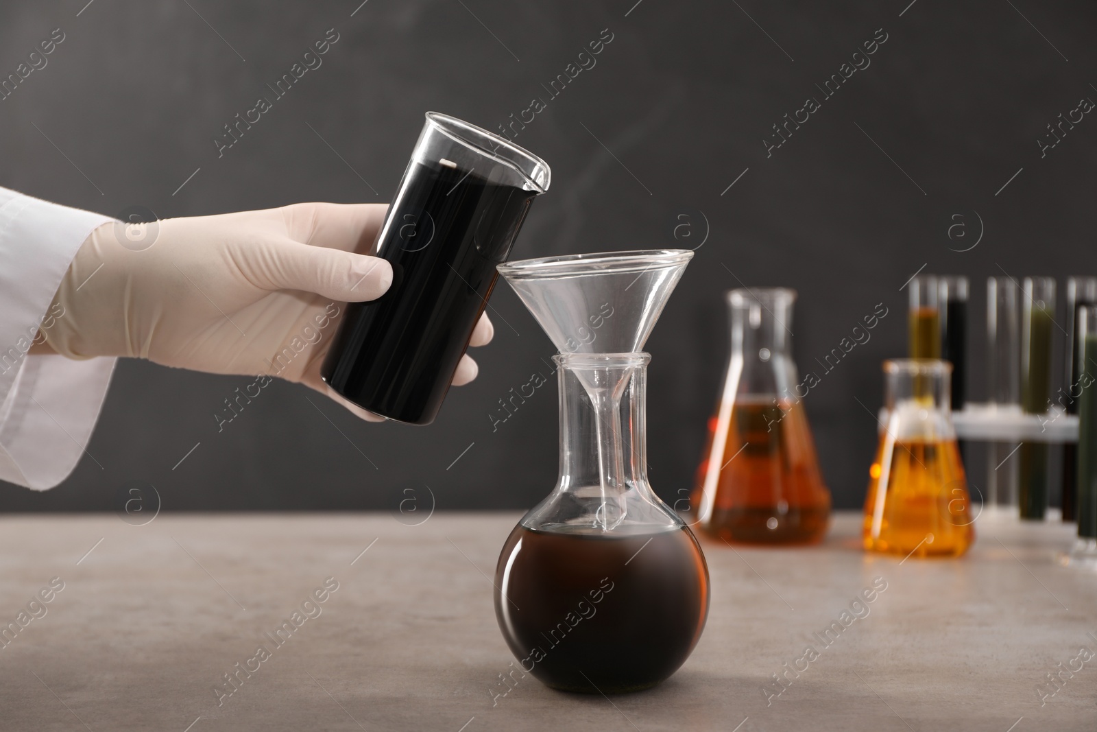 Photo of Woman pouring black crude oil from beaker into flask at grey table, closeup