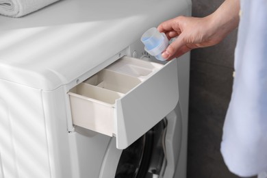 Photo of Woman pouring fabric softener from cap into washing machine near grey wall, closeup