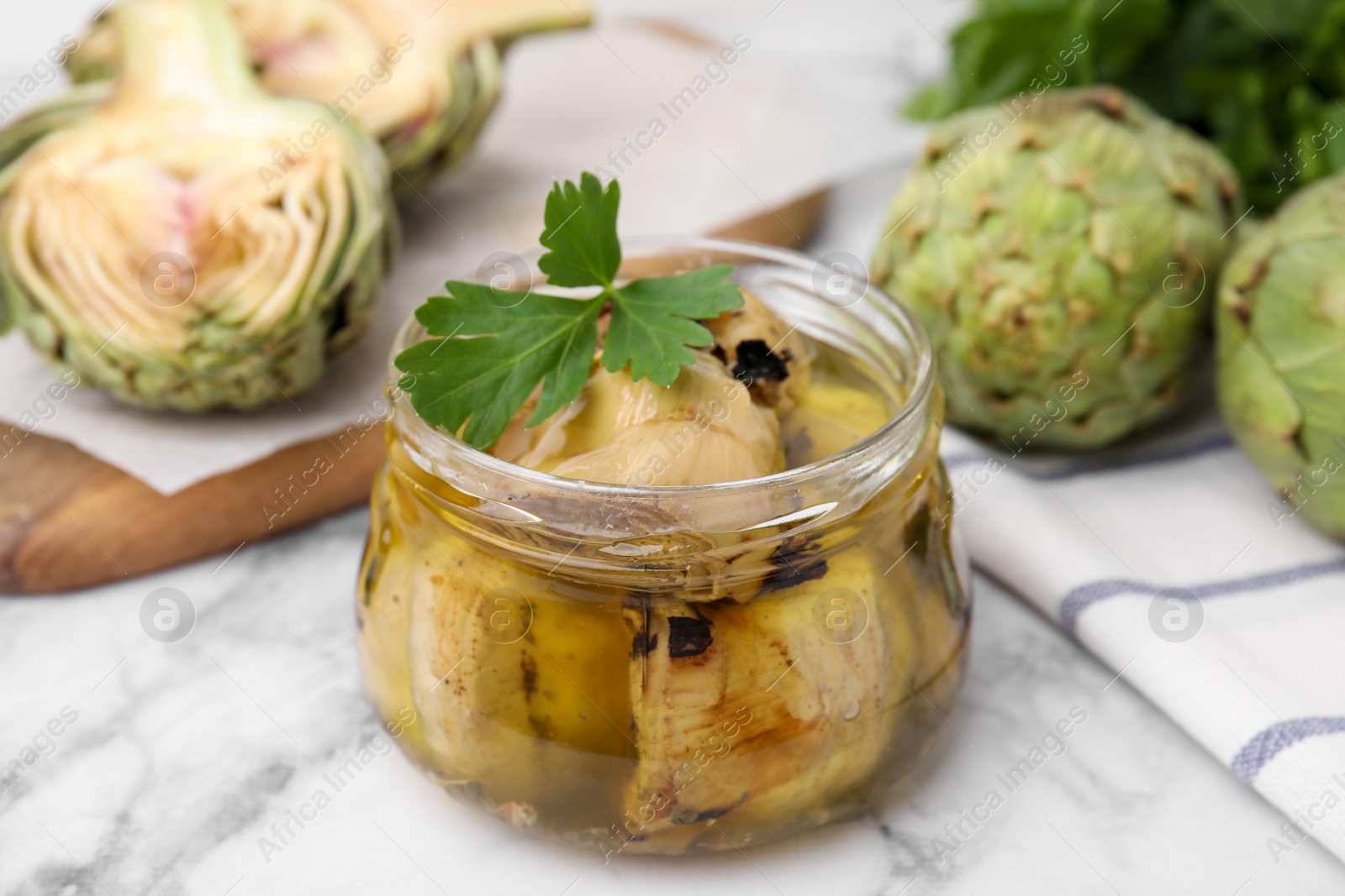 Photo of Jar of delicious artichokes pickled in olive oil and fresh vegetables on white marble table