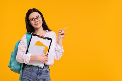 Photo of Smiling student with notebooks and clipboard pointing at something on yellow background. Space for text