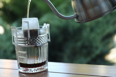 Photo of Pouring hot water into glass with drip coffee bag from kettle on wooden table, closeup