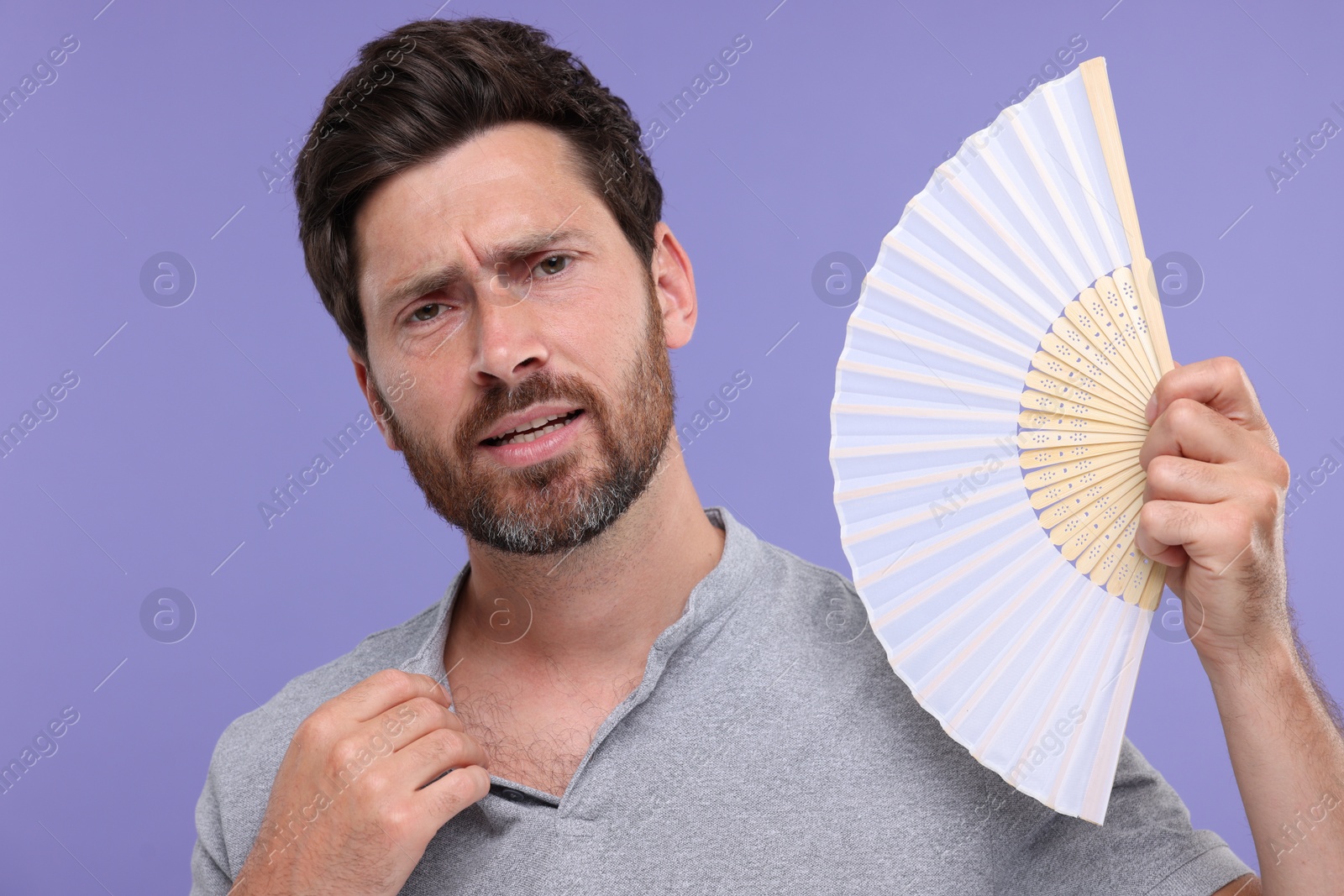 Photo of Unhappy man with hand fan suffering from heat on purple background