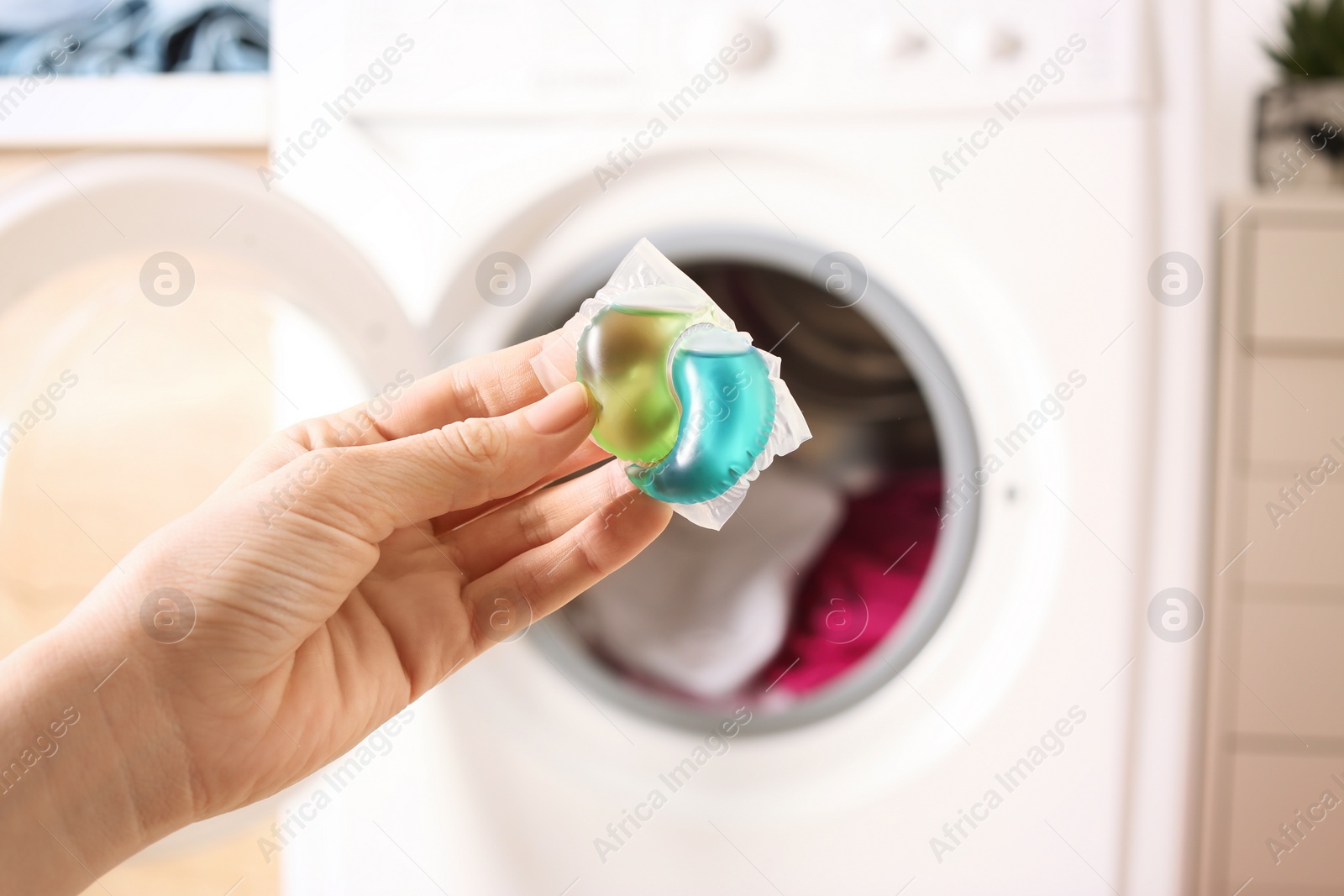 Photo of Woman holding laundry detergent capsule near washing machine indoors, closeup