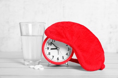 Red sleep mask, glass of water, pills and alarm clock on white wooden table