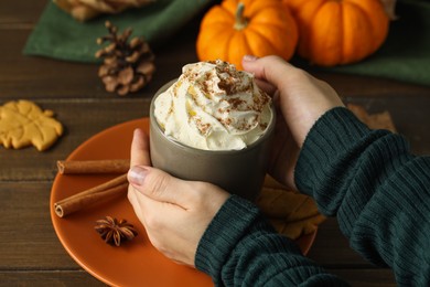 Photo of Woman holding cup of tasty pumpkin spice latte with whipped cream at wooden table, closeup