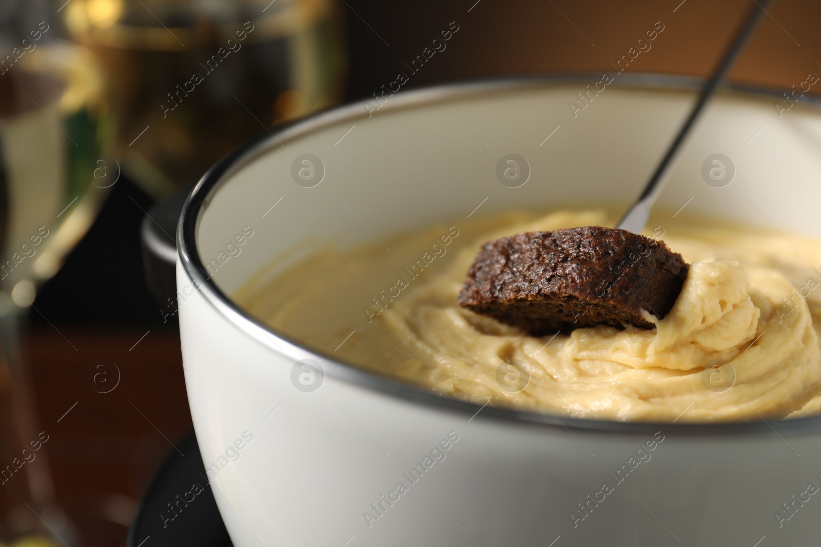 Photo of Dipping piece of bread into fondue pot with melted cheese on dark background, closeup