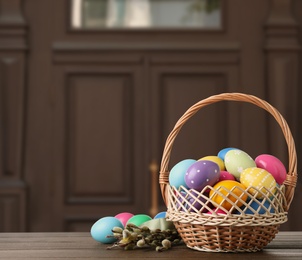 Image of Wicker basket with bright painted Easter eggs on wooden table indoors, space for text 