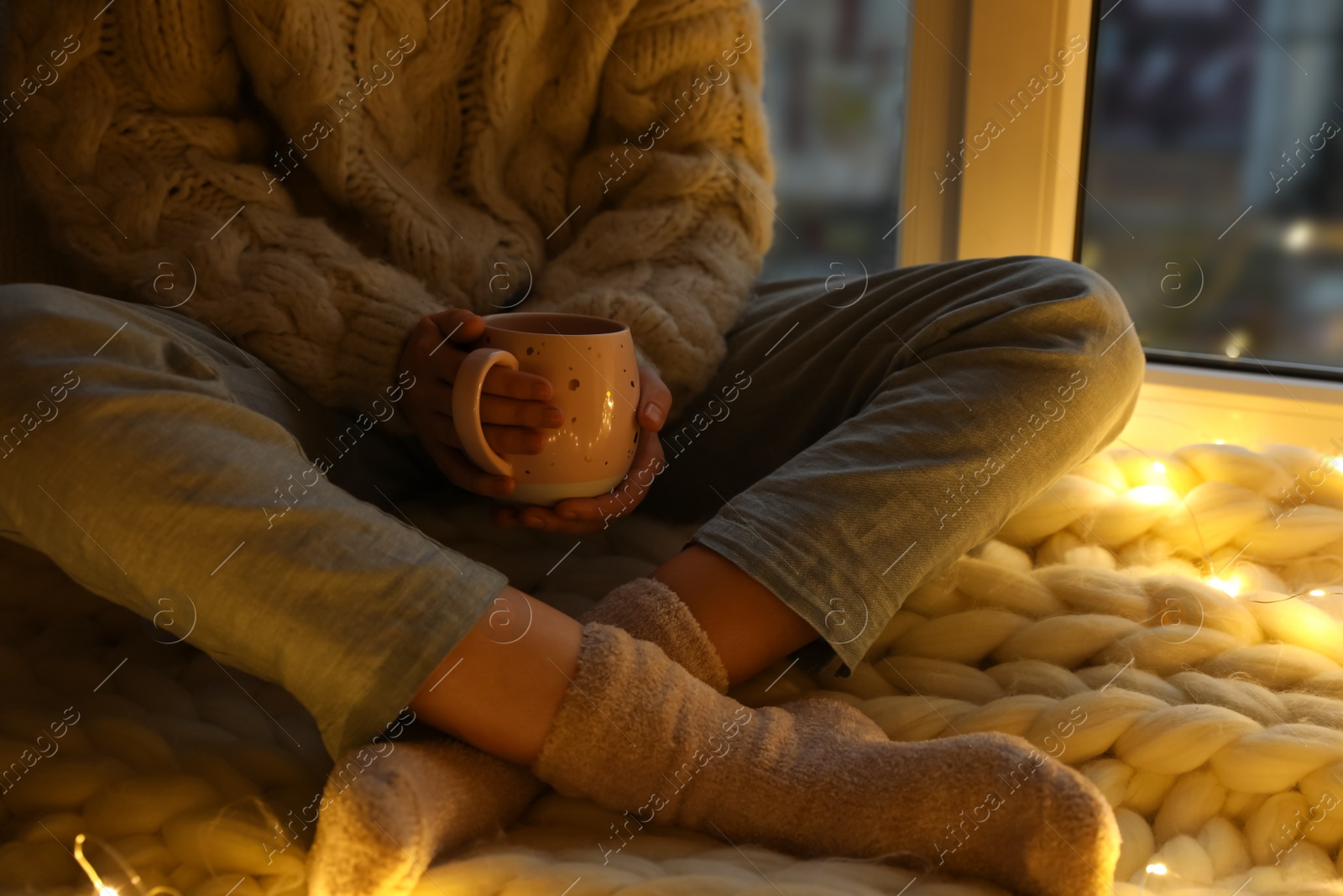 Photo of Woman with drink sitting on soft plaid near window in evening, closeup