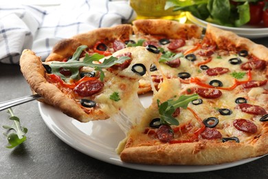 Taking piece of tasty pizza with smoked sausages, olives, pepper and parsley at gray table, closeup