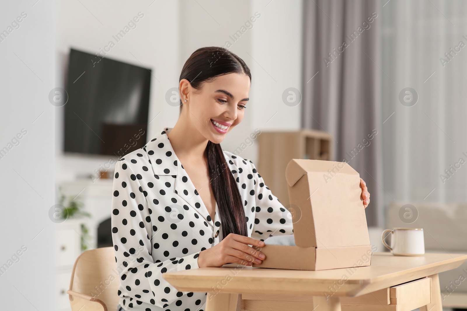 Photo of Happy young woman opening parcel at table indoors. Internet shopping