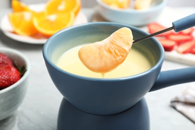 Photo of Dipping tangerine segment into pot with white chocolate fondue on table, closeup