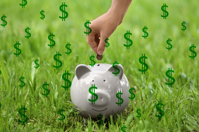 Woman putting coin into piggy bank on green grass outdoors surrounded by dollar signs, closeup