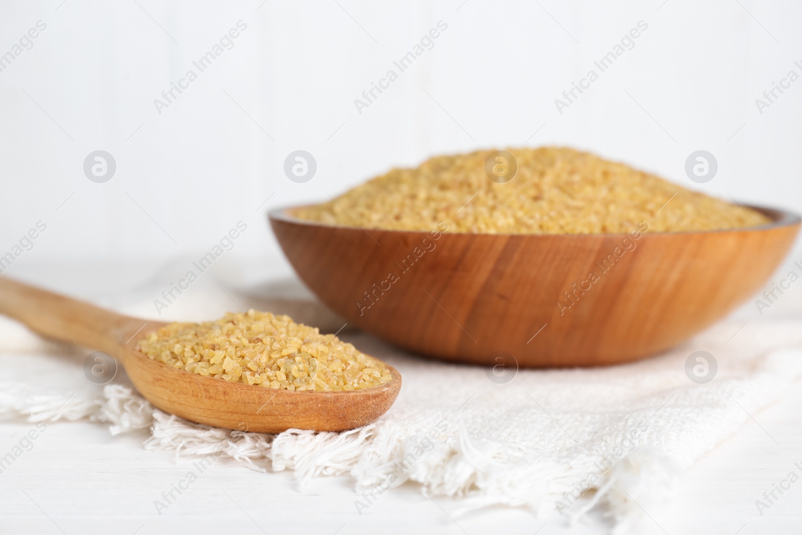 Photo of Napkin, spoon and bowl with uncooked bulgur on white wooden table, closeup