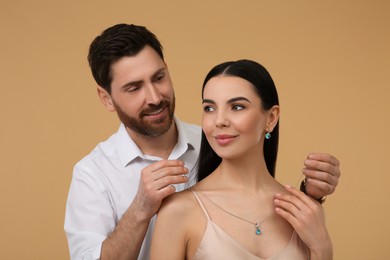 Man putting elegant necklace on beautiful woman against beige background