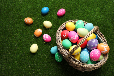 Photo of Colorful Easter eggs in basket on green grass, above view