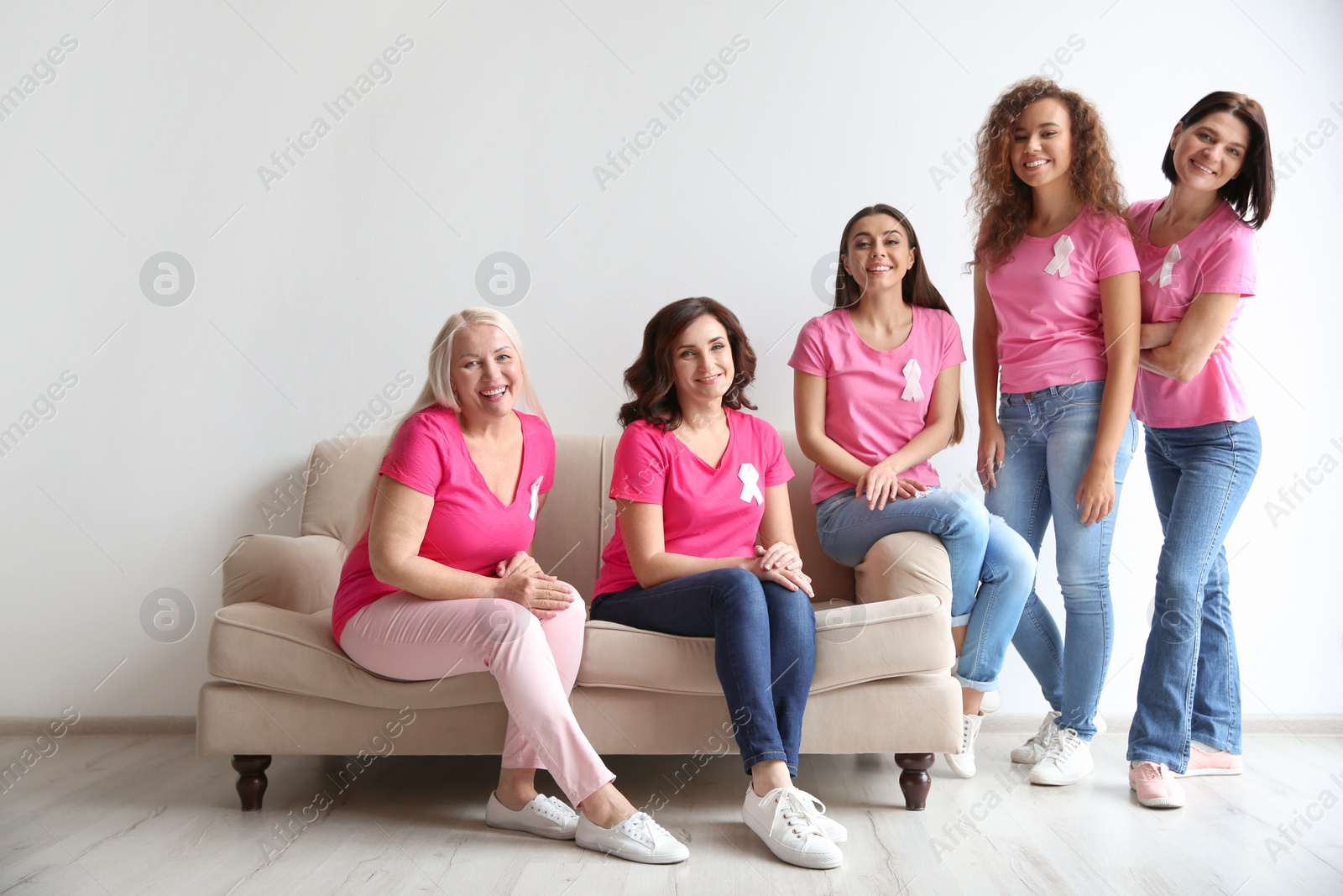 Photo of Group of women with silk ribbons on sofa near light wall. Breast cancer awareness concept