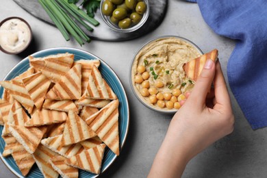 Woman dipping pita chip into hummus at grey table, top view