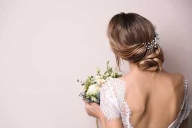 Young bride with elegant hairstyle holding wedding bouquet on beige background, back view. Space for text