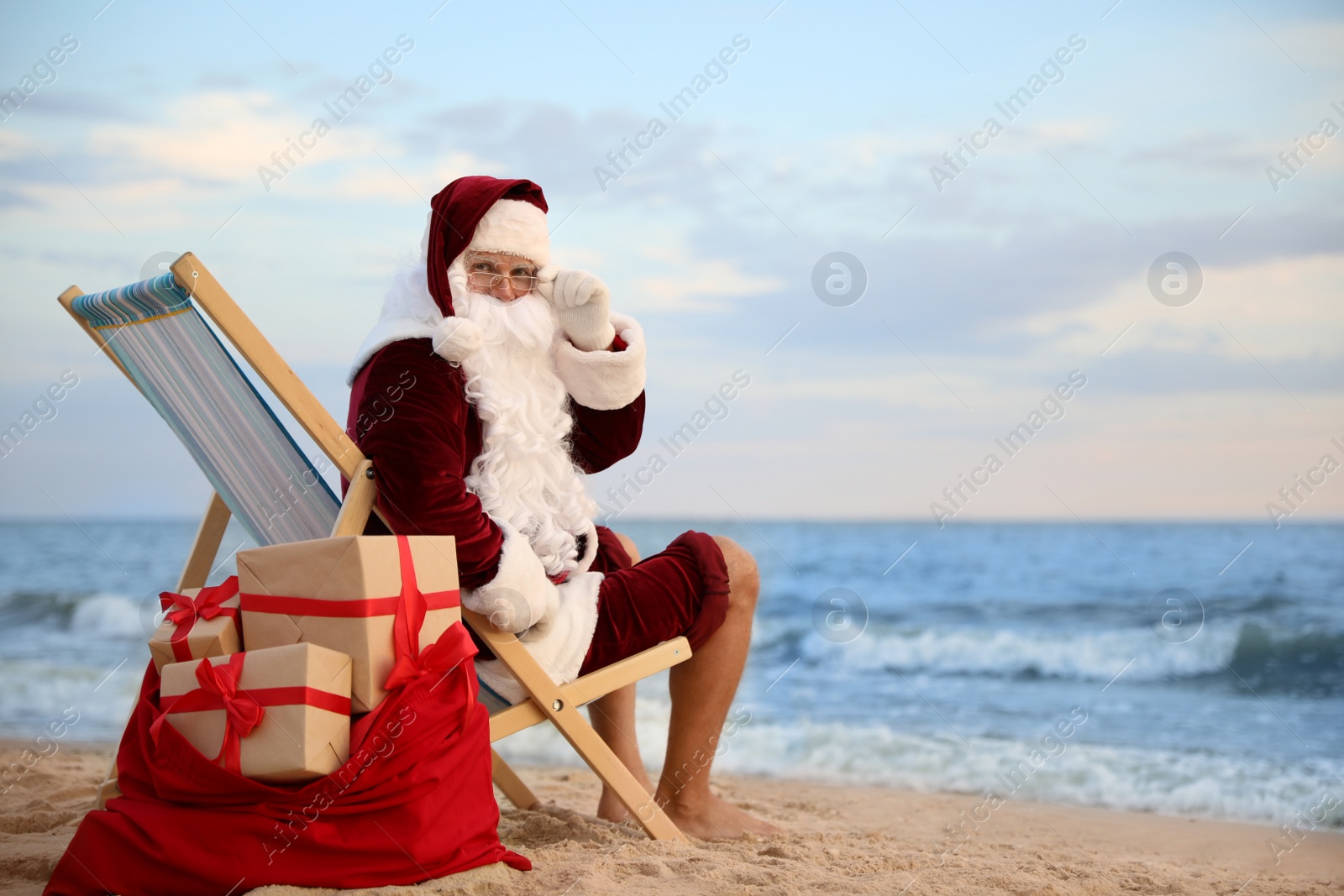 Photo of Santa Claus with bag of presents relaxing in chair on beach, space for text. Christmas vacation