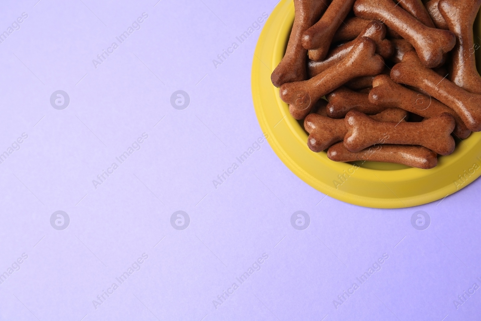 Photo of Yellow bowl with bone shaped dog cookies on purple background, above view. Space for text
