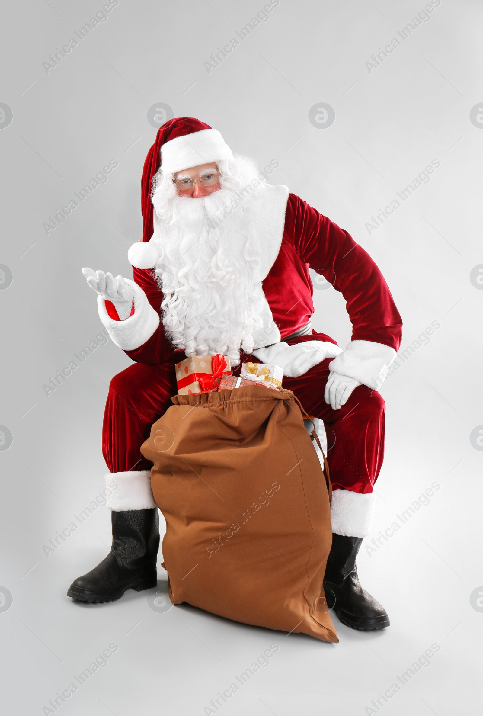 Photo of Santa Claus sitting near sack with gift boxes on light grey background