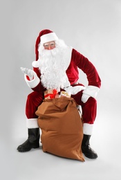 Santa Claus sitting near sack with gift boxes on light grey background