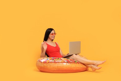 Photo of Happy young woman with beautiful suntan using laptop on inflatable ring against orange background